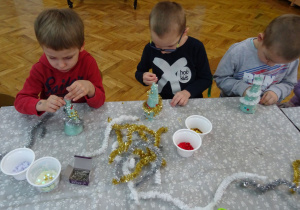 Dzieci siedzą przy stolikach i ozdabiają świątecznymi ozdobami styropianowe choinki.